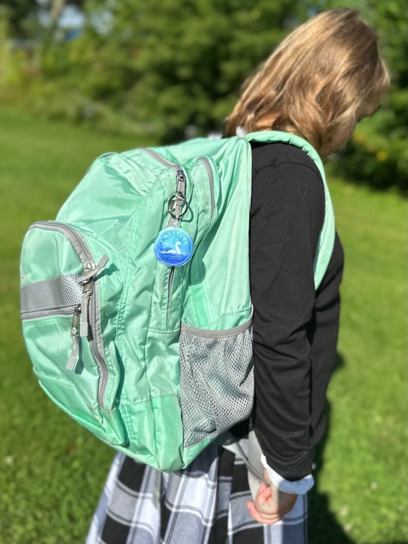 Puede incluir: Una mochila verde claro con un bolsillo de malla gris y una cremallera plateada. Un llavero redondo con un dise&ntilde;o azul y blanco est&aacute; unido al tirador de la cremallera.