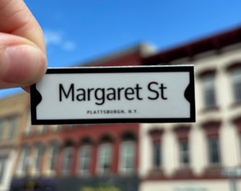 May include: A white sticker with black text that reads "Margaret St. Plattsburgh, N.Y." The sticker is being held up in front of a brick building.