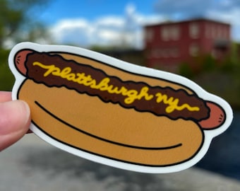May include: A yellow and brown cartoon hot dog sticker with the text "Plattsburgh NY" on top. The sticker is held in a hand against a blurry background of a red building and a blue sky with white clouds.
