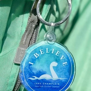 Puede incluir: Un llavero azul y verde con un dise&ntilde;o de acuarela de un monstruo de lago. El llavero dice "I Believe" y "Lake Champlain New York-Quebec-Vermont".