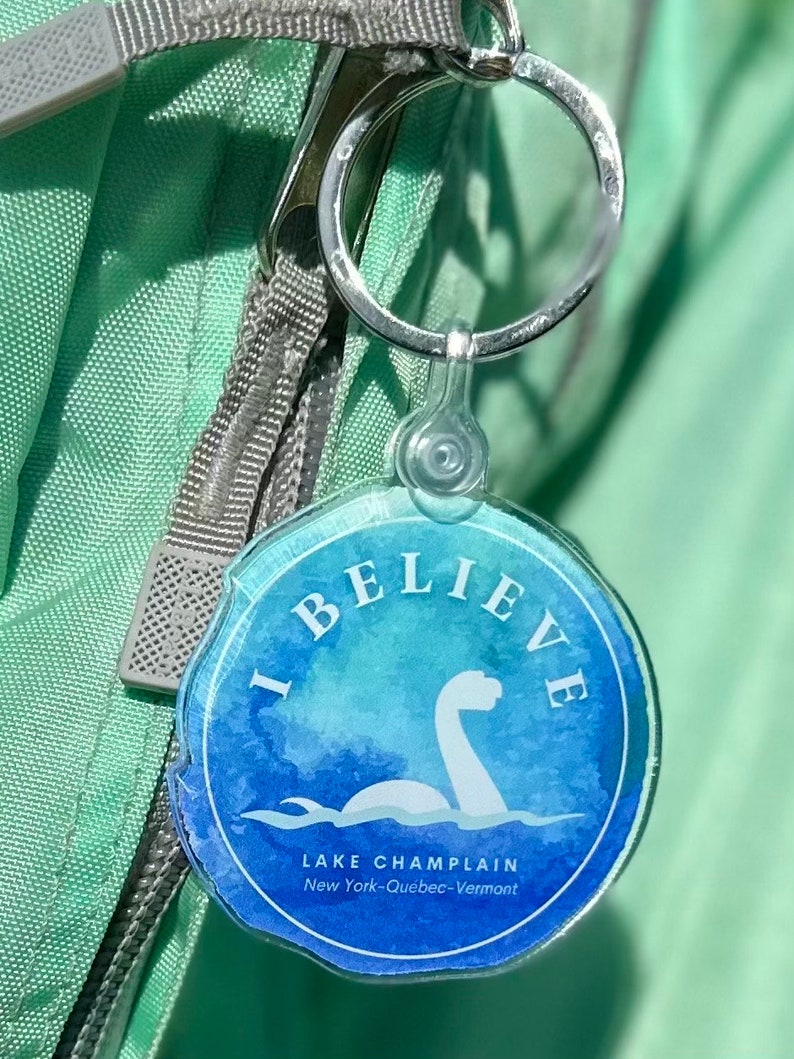 Puede incluir: Un llavero azul y verde con un dise&ntilde;o de acuarela de un monstruo de lago. El llavero dice "I Believe" y "Lake Champlain New York-Quebec-Vermont".