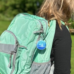 Puede incluir: Una mochila verde claro con una cremallera plateada y un bolsillo de malla. Un llavero redondo con una imagen azul y blanca de un monstruo marino y el texto "I Believe" est&aacute; unido a la cremallera.