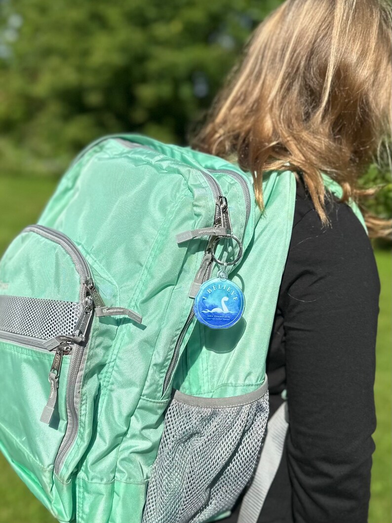 Puede incluir: Una mochila verde claro con una cremallera plateada y un bolsillo de malla. Un llavero redondo con una imagen azul y blanca de un monstruo marino y el texto "I Believe" est&aacute; unido a la cremallera.