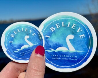 May include: Two round stickers with a watercolor design. The stickers feature a white depiction of a lake monster emerging from blue water, with the text "I BELIEVE" and "LAKE CHAMPLAIN New York-Québec-Vermont".