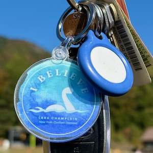 Puede incluir: Un llavero redondo azul y blanco con el texto "I BELIEVE" y "LAKE CHAMPLAIN New York-Quebec-Vermont". El llavero presenta una ilustraci&oacute;n de estilo acuarela de una criatura m&iacute;tica en el agua.
