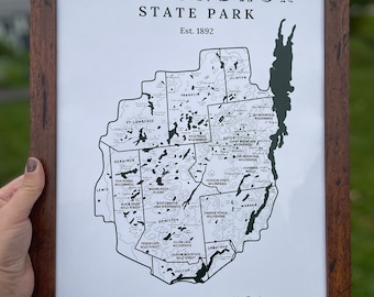 May include: A framed black and white map of Adirondack State Park in New York, with the text "Adirondack State Park Est. 1892" at the top. The map shows the different wilderness areas within the park, including the High Peaks Wilderness, the Giant Mountain Wilderness, and the Jay Mountain Wilderness. The map also includes the coordinates "44.5412°N, 74.6498°W".