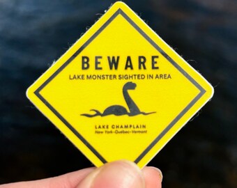 May include: Yellow diamond-shaped warning sign with black text that reads "BEWARE LAKE MONSTER SIGHTED IN AREA". A black silhouette of a sea monster is in the center of the sign. Below the monster is the text "LAKE CHAMPLAIN New York-Quebec-Vermont".