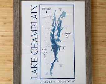 May include: A blue and white map of Lake Champlain with the coordinates 44.5866°N 73.3801°W. The map shows the lake's outline, depth contours, and major cities and towns along its shores. The map is framed in a gray wood frame.