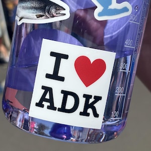 May include: A translucent purple plastic water bottle featuring a white square sticker with the black text I and ADK separated by a red heart. Above this, a sticker depicts a silver and grey fish, alongside a light blue shape and measurement markings.