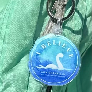 Puede incluir: Un llavero azul y blanco con el texto "I Believe" y "Lake Champlain" con una ilustraci&oacute;n de estilo acuarela de un monstruo marino. El llavero est&aacute; unido a una bolsa verde.