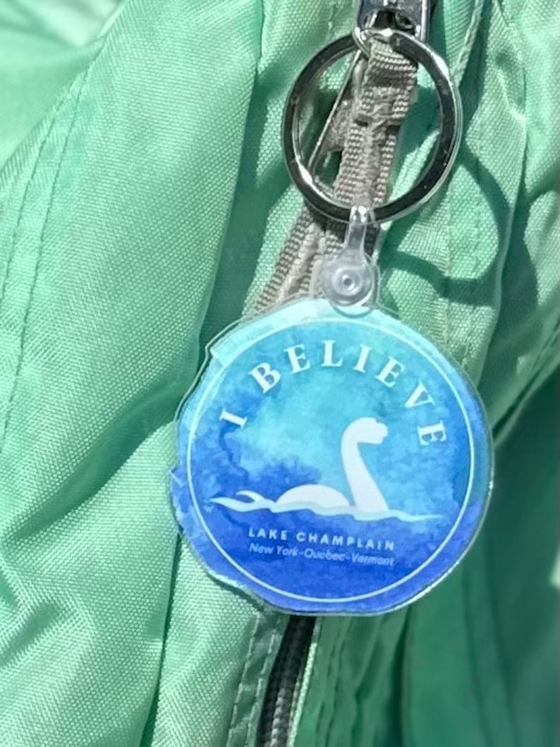 Puede incluir: Un llavero azul y blanco con el texto "I Believe" y "Lake Champlain" con una ilustraci&oacute;n de estilo acuarela de un monstruo marino. El llavero est&aacute; unido a una bolsa verde.
