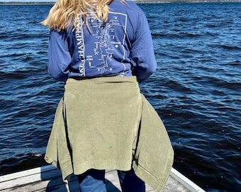 May include: A person wearing a blue long-sleeve shirt with a white graphic of Lake Champlain on the back. The person is standing on a wooden dock overlooking a body of water.