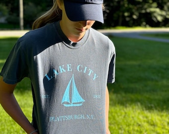 May include: A person wearing a navy blue baseball cap with the number "518" in white and a gray t-shirt. The t-shirt has a light blue sailboat graphic and the words "Lake City Plattsburgh, N.Y."