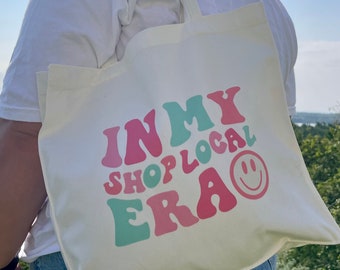 May include: A white canvas tote bag with the text "In My Shop Local Era" in pink, green, and blue letters with a smiley face.