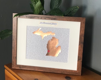 May include: A framed print of the state of Michigan with a gold foil outline and a red heart in the center. The print is surrounded by a white mat and a brown wooden frame. The text "A Thousand Years" is printed at the top of the frame.