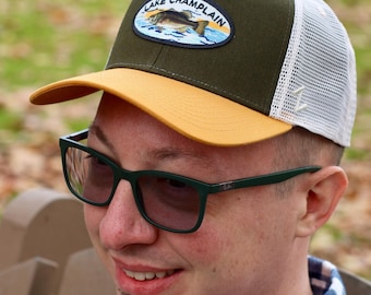 May include: A green and yellow baseball cap with a white mesh back. The cap features a patch with the words "Lake Champlain" and an image of a fish. The person is wearing green-framed glasses.