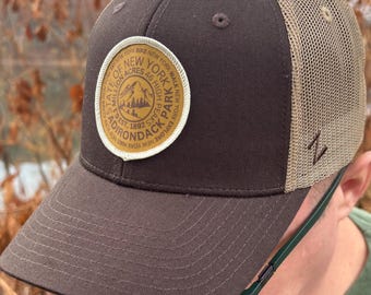 May include: A brown and tan trucker hat with a circular patch featuring text that reads "State of New York Adirondack Park". The hat has a curved brim and a mesh back. The patch includes a mountain graphic.