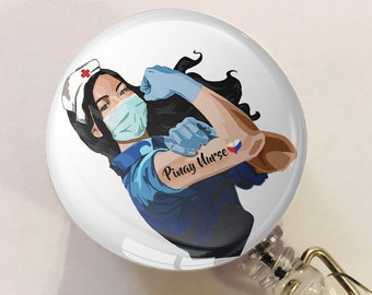 May include: A white round badge holder with a retractable cord and a gold clip. The badge features a cartoon illustration of a woman wearing a blue uniform, a face mask, and a red cross on her hat. The woman has a muscular arm with a tattoo of a heart with the Philippine flag. The text "Pinay Nurse" is written on the badge.