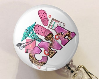 May include: A white retractable badge holder with a pink and white round badge featuring a pink and white medical caduceus, a pink and green pair of clogs, a clipboard with a pink and white checklist, and the text "CNA" in pink with a leopard print background.