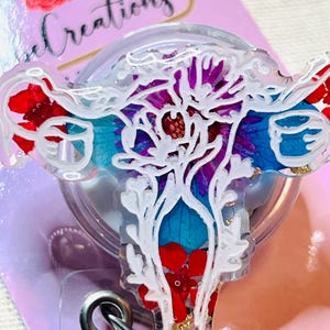 May include: A clear badge reel with a floral design. The design features red, blue, and purple flowers with white accents. The badge reel has a silver metal clip and a retractable cord. The background is a light pink color.