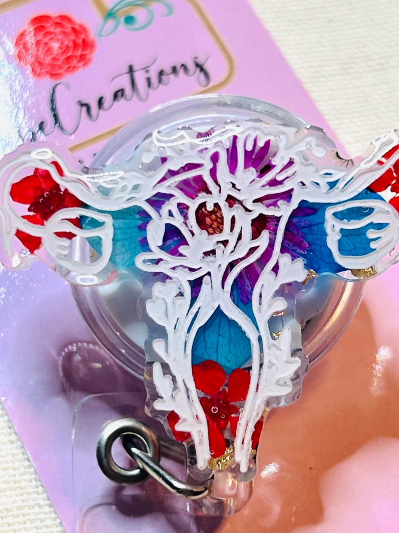 May include: A clear badge reel with a floral design. The design features red, blue, and purple flowers with white accents. The badge reel has a silver metal clip and a retractable cord. The background is a light pink color.