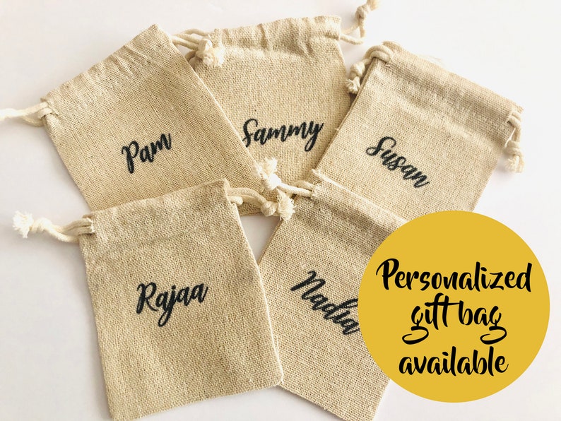 May include: Five beige burlap gift bags with drawstring closures, each personalized with a name in black script: Pam, Sammy, Susan, Rajaa, and Nadia. A yellow circle states "Personalized gift bag available."