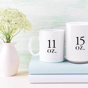 May include: Two white ceramic mugs with black text. The smaller mug says "11 oz." and the larger mug says "15 oz.". The mugs are sitting on top of two light blue books.