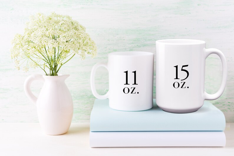 May include: Two white ceramic mugs with black text. The smaller mug says "11 oz." and the larger mug says "15 oz.". The mugs are sitting on top of two light blue books.