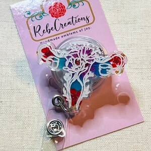 May include: A badge reel featuring a colorful floral design in the shape of a uterus. The badge reel has a clear plastic covering and a metal clip. The backing card is pink with the text "Rebel Creations" and "handmade emblems of joy."