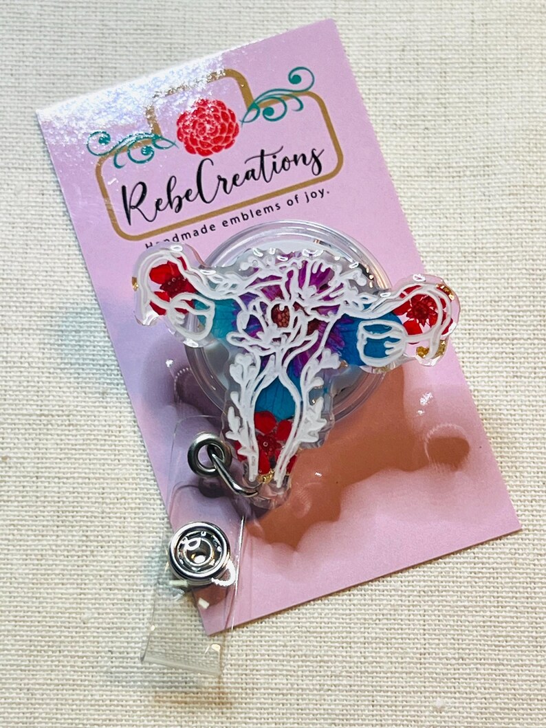 May include: A badge reel featuring a colorful floral design in the shape of a uterus. The badge reel has a clear plastic covering and a metal clip. The backing card is pink with the text "Rebel Creations" and "handmade emblems of joy."