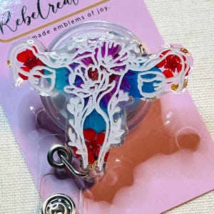 May include: A clear badge reel with a white floral uterus design, filled with red, blue, and purple accents. The badge reel has a silver clip and a snap closure. The text "Rebel Creations" is visible.