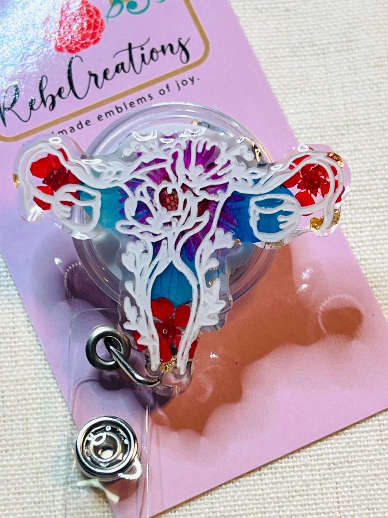 May include: A clear badge reel with a white floral uterus design, filled with red, blue, and purple accents. The badge reel has a silver clip and a snap closure. The text "Rebel Creations" is visible.