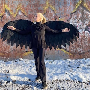 Könnte beinhalten: Eine Person trägt ein schwarzes Hemd mit Stehkragen und eine schwarze Hose mit großen schwarzen gefiederten Flügeln, die hinter ihr ausgebreitet sind. Die Person steht vor einer mit Graffiti besprühten Wand mit Schnee auf dem Boden.