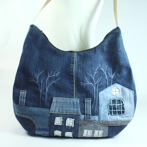 Peut inclure: Un sac à bandoulière en jean avec un motif de paysage. Le sac présente un extérieur en jean bleu avec une sangle beige. Le motif de paysage comprend des arbres brodés et des maisons appliquées dans des tons de bleu et de gris.