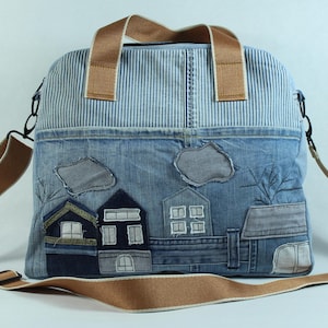 Puede incluir: Bolso tote de mezclilla con una escena de pueblo aplicada. El bolso tiene una parte superior a rayas azules y blancas, asas marrones y una correa ajustable para el hombro. La escena del pueblo incluye casas, nubes y árboles. Medidas: 30 cm x 30 cm.