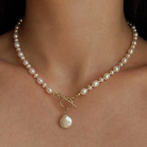 Può includere: Una collana composta da una fila di piccole perle color bianco sporco di forma irregolare. La collana presenta una chiusura a T in metallo dorato posizionata al centro, da cui pende un singolo ciondolo di perla rotonda, piatta e più grande.