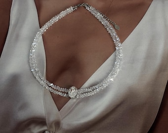 May include: A double-strand necklace composed of clear, faceted crystal beads featuring a central oval-shaped crystal pendant. The necklace is worn against a white satin sleeveless top with a deep V-neckline and a twisted knot detail at the centre.