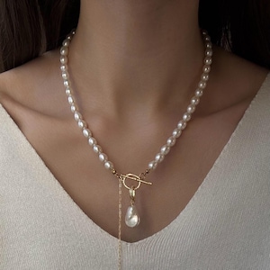 Peut inclure: Un collier composé d'un rang de perles blanches ovales reliées par un fermoir à bascule doré. Une perle unique en forme de goutte plus grande est suspendue à un anneau doré fixé au fermoir, avec une fine chaîne dorée tombant vers le bas.