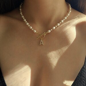 May include: A pearl necklace with a gold clasp and a pendant featuring the letter "A" encrusted with small, clear stones. The necklace is composed of small, round pearls and is worn around the neck.
