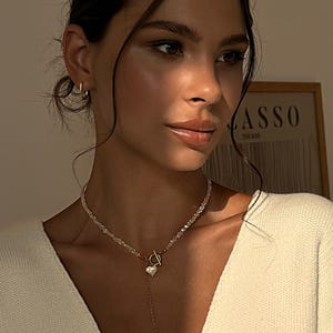 以下が含まれることがあります： ハートのペンダントとビーズチェーンが付いた繊細なネックレス。クリーム色のセーターの上から着用されています。画像は明るく照らされており、ジュエリーの詳細とセーターの柔らかな質感を際立たせています。