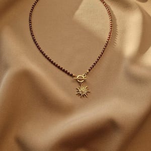 May include: A delicate necklace featuring a strand of small, dark red beads. The necklace has a gold-colored clasp and a sunburst pendant. The jewelry is displayed on a tan fabric background.