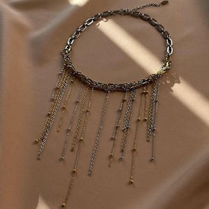Puede incluir: Un collar plateado y dorado con múltiples hilos de delicadas cadenas y pequeñas cuentas. El collar presenta un diseño en capas con un collar de cadena y flecos en cascada, creando un accesorio elegante.