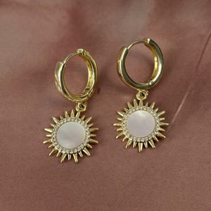 Peut inclure: Une paire de boucles d'oreilles cr&eacute;oles dor&eacute;es avec des pendentifs en forme de soleil. Chaque pendentif pr&eacute;sente une incrustation circulaire en nacre blanche entour&eacute;e de petits cristaux clairs et de rayons dor&eacute;s pointus sur le bord.