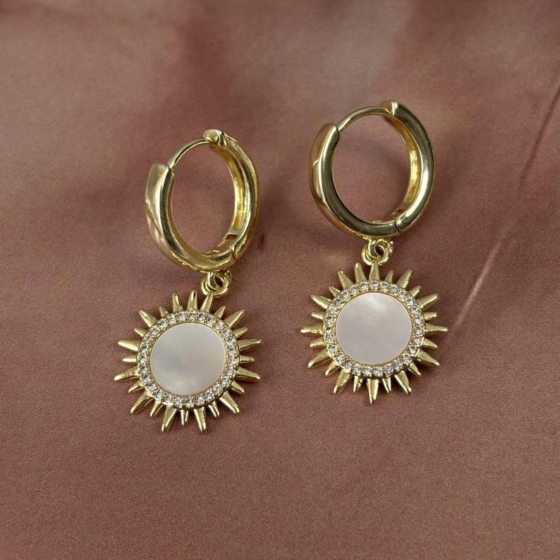 Peut inclure: Une paire de boucles d'oreilles cr&eacute;oles dor&eacute;es avec des pendentifs en forme de soleil. Chaque pendentif pr&eacute;sente une incrustation circulaire en nacre blanche entour&eacute;e de petits cristaux clairs et de rayons dor&eacute;s pointus sur le bord.