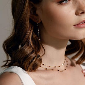 Può includere: Una collana a catena dorata con due strati delicati decorati con piccole perline rosso scuro. Il set di gioielli include un orecchino pendente singolo abbinato con una catena dorata simile e un dettaglio a perla rosso scuro sul lobo.