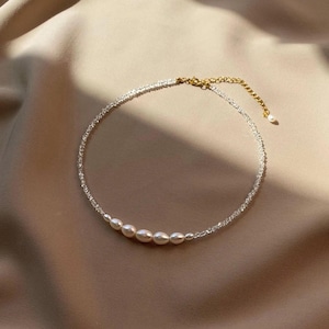 Könnte beinhalten: Zarte Halskette mit einer Reihe kleiner, glänzender Perlen. Die Halskette hat eine silberfarbene Kette mit einem goldfarbenen Verschluss und einer einzelnen Perle am Ende. Der Schmuck wird auf einem neutralfarbenen Stoff präsentiert.