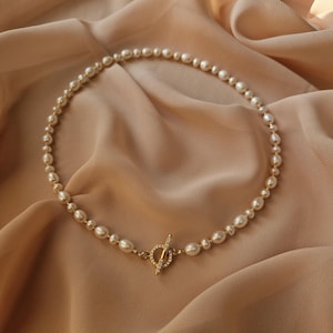 May include: A delicate pearl necklace with a gold clasp. The necklace features a strand of white pearls of varying sizes, connected by a gold chain. The clasp is a gold toggle bar with small crystals. The necklace is displayed on a beige fabric.