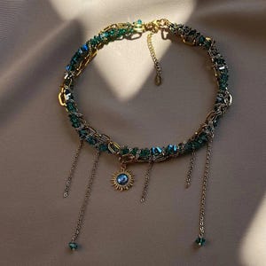 Peut inclure: Un collier doré avec un pendentif soleil et des chaînes pendantes. Le collier présente un design tressé avec des perles de cristal vertes et bleues. Le pendentif soleil a un centre bleu et est entouré de rayons dorés. Le collier est sur un fond neutre.