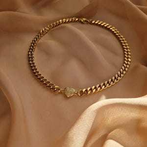 May include: A gold-coloured chain necklace with a heart-shaped pendant. The necklace features a thick curb chain design and a small heart pendant with a textured surface. The jewellery is displayed on a beige fabric background.