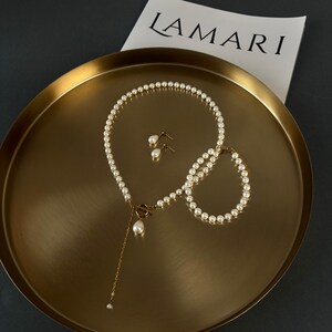 May include: A set of pearl jewellery, including a necklace, bracelet, and earrings, displayed on a gold-coloured tray. The necklace features a gold clasp and a dangling pearl. The earrings are small pearl studs. A paper with "LAMARI" is also on the tray.